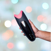 A hand is holding the sleek Maia Piper Multi-Function Rechargeable Male Masturbator by Maia Toys, featuring a stylish black exterior with a pink interior. The background is artfully blurred with multicolored bokeh lights. Buttons on the side and its glossy finish hint at the device's electronic sophistication and 15 vibration suction settings.